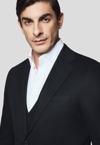 Czarny garnitur na miarę z ostrymi klapami, zestawiony z białą koszulą elegancką. Gładka faktura materiału i subtelne detale szycia.