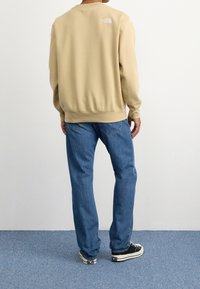 Beige mikina s kulatým výstřihem a ribbovaným lemem, na zadní straně má malé bílé logo. V kombinaci s modrými džíny a černými teniskami.
