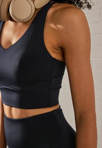 Zwarte sport-bh met brede bandjes, een V-halsontwerp en naadloze textuur, aangevuld met grote beige hoofdtelefoon om de nek.