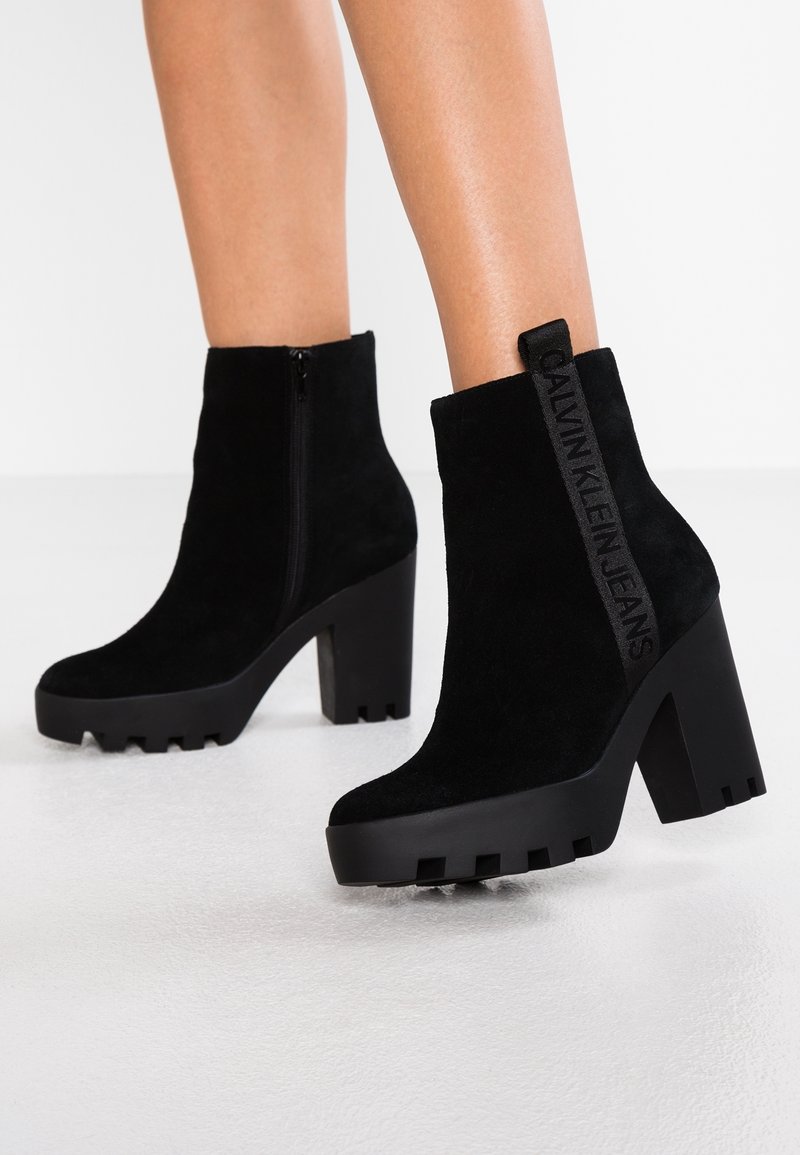 Mustat mokkanahkasaappaat nilkkoihin, joissa on paksu korko, vetoketjukiinnitys ja brändätty silmukka takana. Teksturoitu pohja pidon lisäämiseksi.