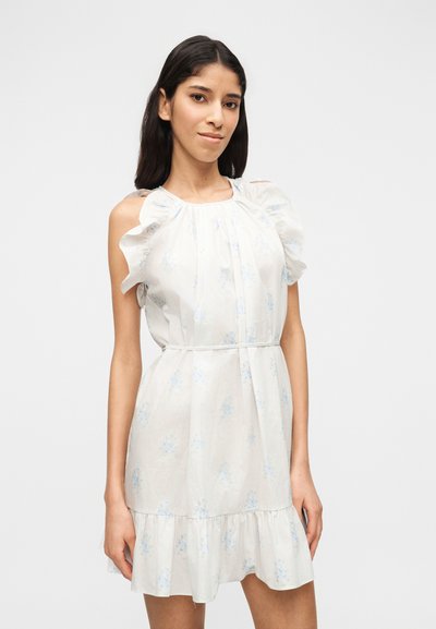 Νεαρή γυναίκα με μακριά σκούρα μαλλιά, ντυμένη με λευκό αμάνικο φόρεμα με βολάν στους ώμους και ανοιχτό μπλε φλοράλ σχέδιο, που στέκεται μπροστά σε απλό φόντο.