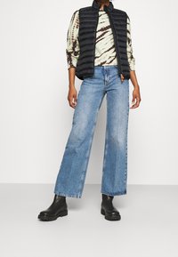 Sort puffer-vest over en sweater med tie-dye mønster, kombineret med vide blå jeans og sorte ankelstøvler, med teksturerede materialer og et strømlinet design.