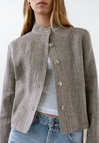 Brown linen jacket with a collar, button closure, and a textured finish. Worn over a white top, paired with light blue denim jeans.