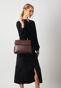 Brown leather handbag with a structured design and gold hardware. Worn by a person in a black top and skirt, showcasing a side slit.