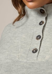 Pull en maille côtelée gris clair avec un col montant et quatre boutons foncés sur le devant. Texture douce avec une coupe décontractée et ajustée.