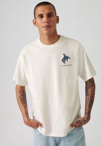 Hvid bomuld t-shirt med en marineblå silhuet af en rytter og "Levi Strauss & Co."-tekst på venstre bryst. Afslappet pasform, korte ærmer.