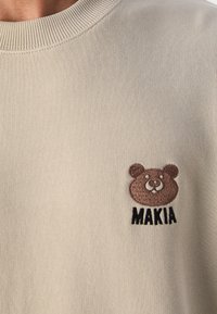 Maglione beige con scollo a costine e logo di orso marrone ricamato. Testo "MAKIA" sotto l'orso in nero. Tessuto morbido e testurizzato.