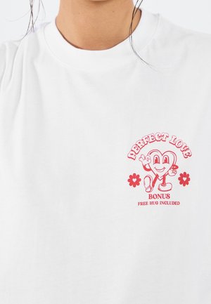 Wit katoenen T-shirt met een rode afbeelding van een glimlachend hartfiguurtje met de tekst "PERFECT LOVE" en "BONUS GRATIS OMHELZING INBEGREPEN."