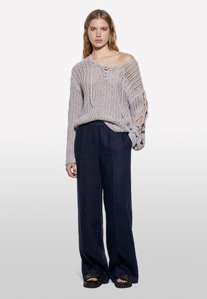Un pull en tricot épais gris clair avec un décolleté à lacets et un design de manches ouvertes, associé à un pantalon ample bleu marine et des sandales noires.