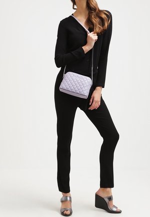 Vadderad lavendelfärgad crossbody-väska med silverkedja; modell bär en svart topp och byxor samt metalliska kilskor.