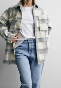 Licht gewatteerd grijs-wit geruit flanellen overhemd met een kraag, twee voorzakken en knopen, gecombineerd met lichtblauwe jeans.