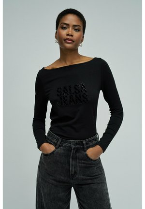 Femme portant un haut noir à manches longues avec le texte "SALSA JEANS" et un jean noir délavé taille haute, mains dans les poches, sur un fond uni.