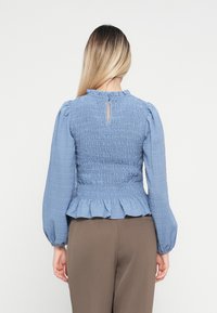 Blå smockbluse med nøglehulslukning, flæser i kanten og voluminøse ærmer. Lavet af letvægts tekstureret stof. Sammen med brune bukser.