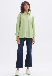 Light green button-up shirt with a front pocket, paired with dark blue flared jeans and white sneakers with green accents.