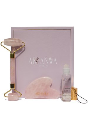 Rouleau facial en quartz rose, outil gua sha et sérum éclat cristal devant une boîte rose Aranwa Skincare.