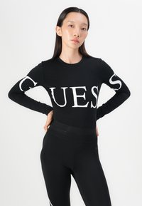 Crna majica dugih rukava s velikim bijelim natpisom "GUESS" na prednjoj strani, u kombinaciji s crnim tajicama visokog struka. Glatka, rastezljiva tkanina.