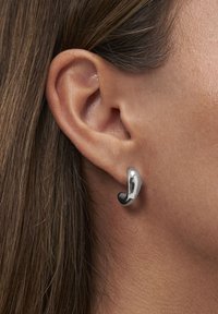 Nahaufnahme eines linken Ohrs mit glattem braunem Haar, das einen kleinen, glänzenden, gebogenen silbernen Ohrring trägt.