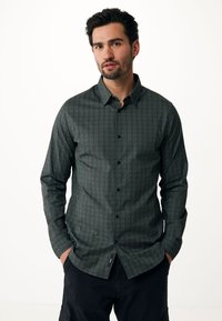 Camicia a maniche lunghe con motivo verde a trama a spina di pesce, bottoni neri. Presenta un orlo curvato e un dettaglio con etichetta laterale.