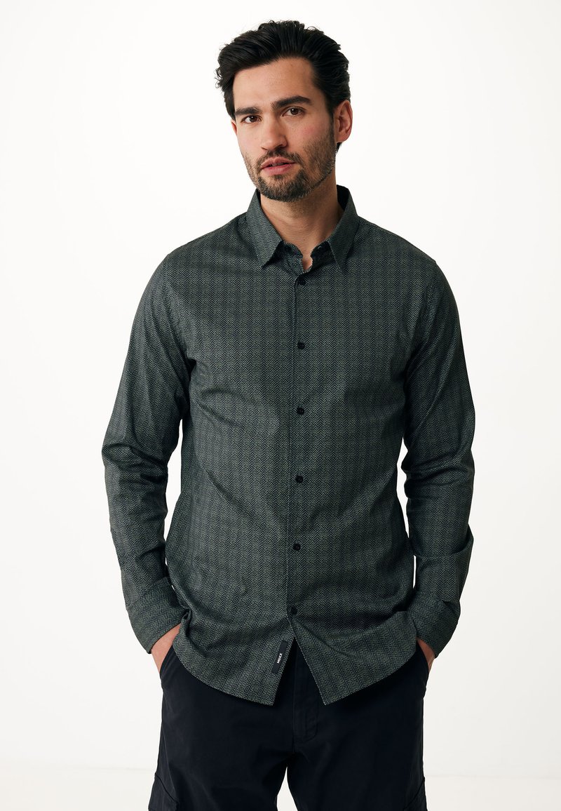 Camicia a maniche lunghe con motivo verde a trama a spina di pesce, bottoni neri. Presenta un orlo curvato e un dettaglio con etichetta laterale.