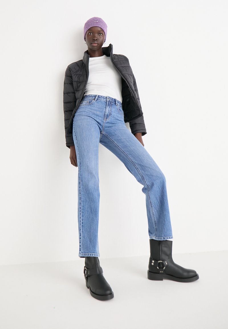 Chaqueta acolchada negra sobre una camiseta blanca, pantalones vaqueros de pierna recta azules y botas negras hasta el tobillo con hebillas; gorro de lana morado.