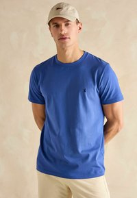 Niebieska bawełniana koszulka z krótkim rękawem, okrągłym dekoltem i małym logo na piersi, noszona z jasnym czapką. Gładka faktura.