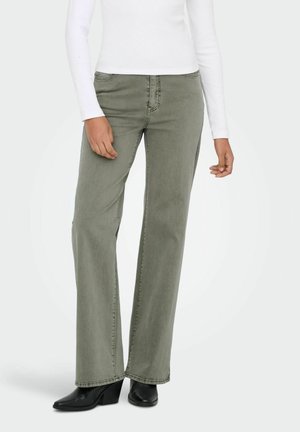 Nő, aki olíva zöld bőszárú farmernadrágot, fekete magas sarkú csizmát és fehér, hosszú ujjú felsőt visel, és sima háttér előtt áll.