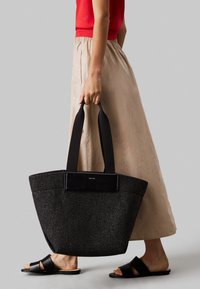 Borsa tote nera intrecciata con superficie texture, dotata di una tracolla rettangolare nera e un'accattivante accentuazione del logo. Portata a mano insieme a una gonna di colore chiaro.