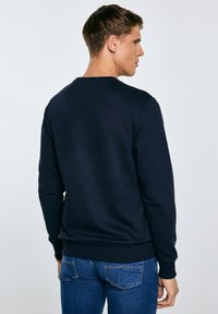 Jaunas vyras nugara, vilkintis paprastą tamsiai mėlyną džemperį ir mėlynas džinsus, priešais šviesiai pilką foną.