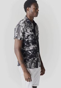 Homme debout de profil portant une chemise à manches courtes à motifs abstraits noir et blanc et un short blanc devant un fond uni.