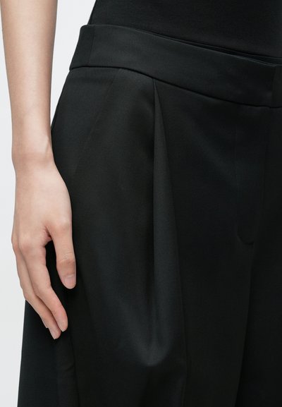 Pantalon noir plissé en tissu lisse, avec une large ceinture et une coupe décontractée offrant un drapé notable et une légère brillance.