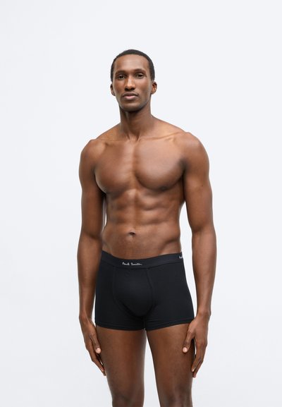 Schwarze Boxer-Shorts aus weichem Material mit enger Passform. Verfügt über einen gebrandeten Bund mit weißem Text. Das Modell steht vor einem einfarbigen Hintergrund.