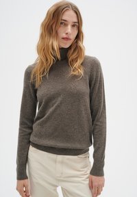 Brun stickad polotröja med långa ärmar och ribbade muddar, med en mjuk textur och en något lös passform. Bärs med ljusa byxor.