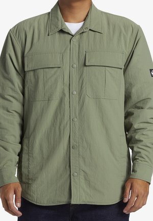 Mężczyzna noszący jasnooliwkową koszulę zapinaną na guziki, z dwoma patkami na piersi i długimi rękawami.