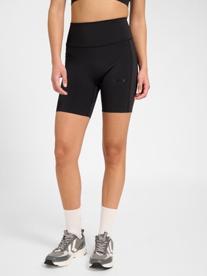 Femme portant un short cycliste noir taille haute, des chaussettes blanches et des chaussures de sport grises, debout sur un fond uni.