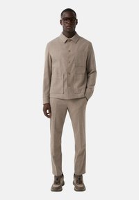 Beige uldblandingsjakke med knaplukning, krave og brystlomme, parret med matchende bukser og strukturerede brune sneakers.