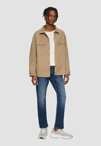 Veste en coton beige avec deux poches avant, portée sur une chemise blanche, associée à un jean bleu et des baskets grises.