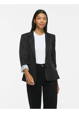 Donna con lunghi capelli scuri che indossa un blazer nero sopra una camicia bianca e pantaloni neri, in piedi davanti a uno sfondo bianco uniforme.