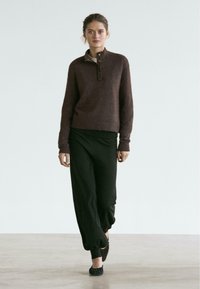 Brauner Wollpullover mit hohem Kragen und Drei-Knopf-Leiste, kombiniert mit schwarzen, locker sitzenden Hosen mit gerafften Bündchen.