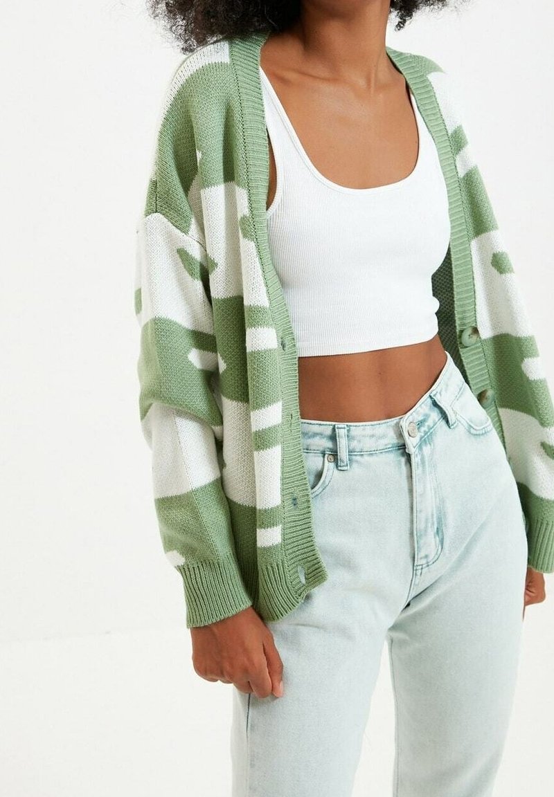 Femme portant un cardigan à motifs vert clair et blanc par-dessus un débardeur court blanc et un jean taille haute bleu clair.