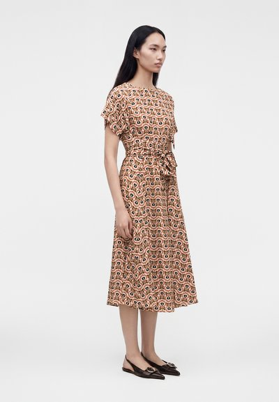 Femme debout portant une robe marron à motifs longueur genou avec manches courtes et taille nouée, associée à des chaussures plates pointues noires.