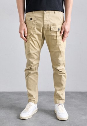 Pantalones cargo beige con múltiples bolsillos, cierre de botón y un diseño de pierna ajustada, combinados con zapatillas blancas sobre un suelo gris.