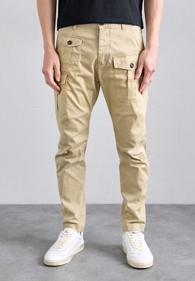Pantalones cargo beige con múltiples bolsillos, cierre de botón y un diseño de pierna ajustada, combinados con zapatillas blancas sobre un suelo gris.