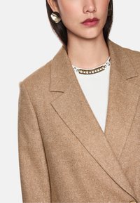 Brauner Wollmischung-Sakko mit strukturiertem Finish, mit Reverskragen, kombiniert mit einem weißen Oberteil mit einem mit Juwelen besetzten Kragen. Runde metallische Ohrringe.