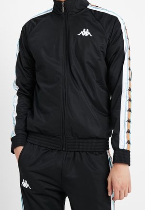 Czarny dres Kappa z kurtką na pełny zamek i pasującymi spodniami, ozdobiony białą i pomarańczową taśmą z logo wzdłuż rękawów i nogawek.