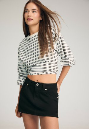 Jeune femme aux longs cheveux bruns portant un crop top rayé blanc et noir et une mini-jupe en jean noire, debout avec une main sur la hanche.