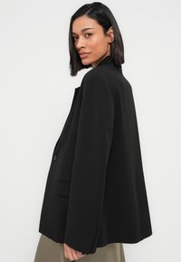 Γυναίκα με ίσια σκούρα μαλλιά που φοράει χαλαρό μαύρο σακάκι και μπεζ παντελόνι, κοιτάζοντας πάνω από τον ώμο της σε λευκό απλό φόντο.