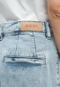 Ανοιχτό μπλε τζιν με δερμάτινη ετικέτα που γράφει "JOOP!" πάνω από μια οριζόντια πίσω τσέπη, συνδυασμένο με λευκό τοπ.