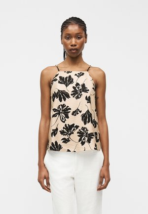 Γυναίκα με πλεξούδες φοράει μπεζ αμάνικη μπλούζα με μαύρο λουλουδένιο σχέδιο και λευκό παντελόνι, στέκεται μπροστά σε απλό φωτεινό φόντο.