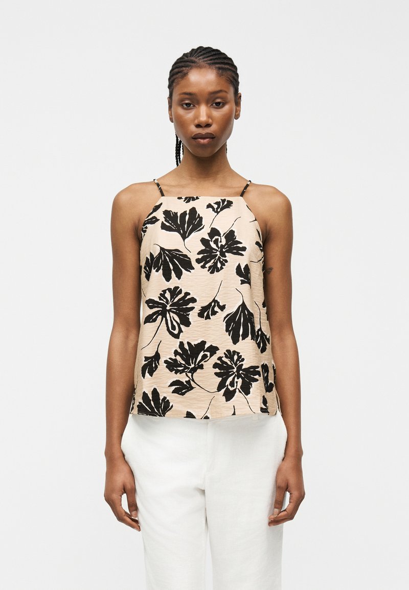 Γυναίκα με πλεξούδες φοράει μπεζ αμάνικη μπλούζα με μαύρο λουλουδένιο σχέδιο και λευκό παντελόνι, στέκεται μπροστά σε απλό φωτεινό φόντο.