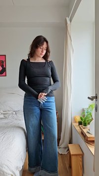 Top nero a maniche lunghe con scollatura quadrata, abbinato a jeans larghi blu chiaro con orli sfrangiati. La luce naturale illumina la scena.